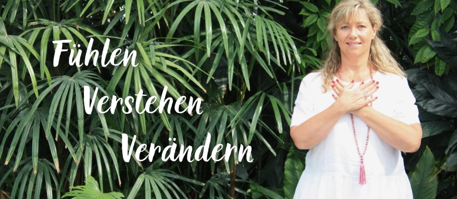 Olga Ahl mit der Hand auf dem Herzen vor grünen Pflanzen – Bewusstseinsarbeit und persönliche Begleitung